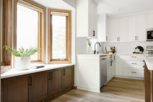 A bay window lets light into a mostly white kitchen. Small wooden modern baseboard accent.